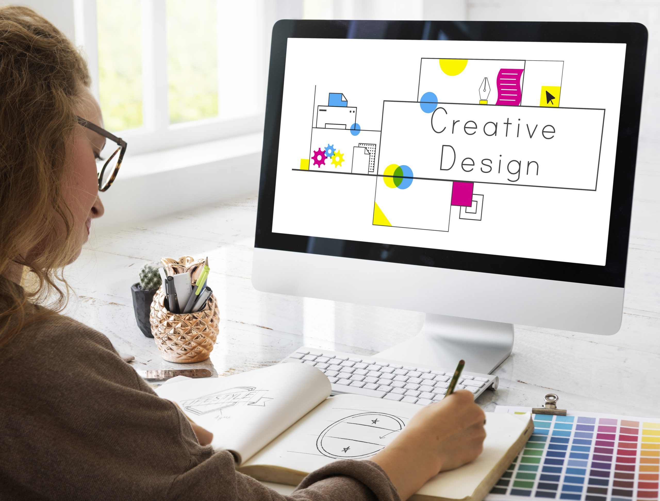 Designer sketching in a notebook while a computer screen shows a 'Creative Design' layout with colorful shapes and text, at a bright desk.