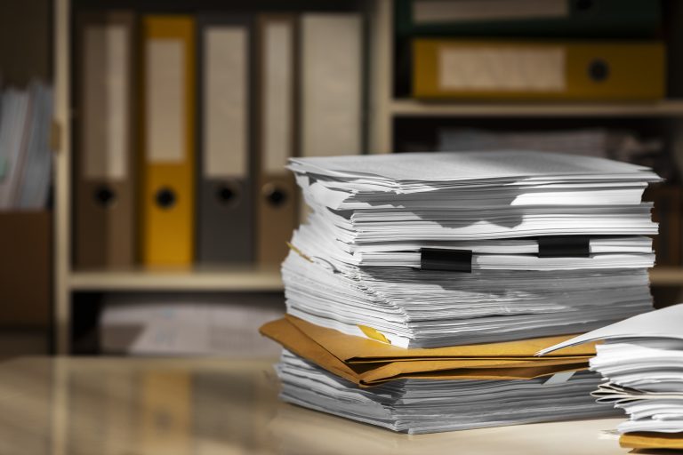 A tall pile of loose papers and manila folders on a desk with filing boxes on a blurred office shelf behind it.