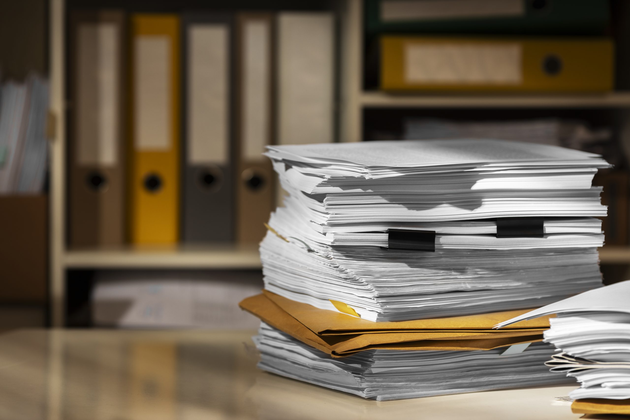 A tall pile of loose papers and manila folders on a desk with filing boxes on a blurred office shelf behind it.